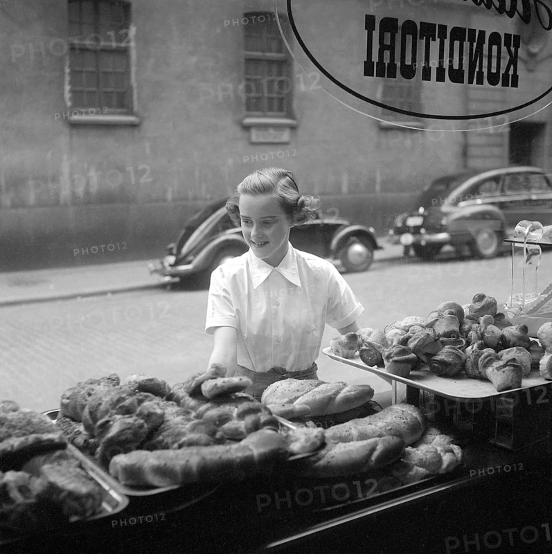 In the 1950s. A woman is pictured in the street and at the storefront of a bakery. She looks as if she is deciding what to buy and looks tempted by the good selection of sweet bread and buns. Sweden 1951 Kristoffersson ref BD91-5