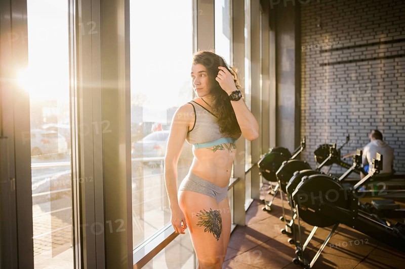 Young caucasian sporty woman with curly long hair poses in sportswear near a panorman window in a fitness club. Young female athlete model with beautiful body stands in the gym with a tattoo