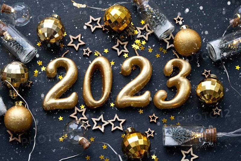 The golden figures 2023 made of candles on a black stone slate background are decorated with a festive decor of stars, sequins, fir branches, balls and garlands. Greeting card, happy New Year