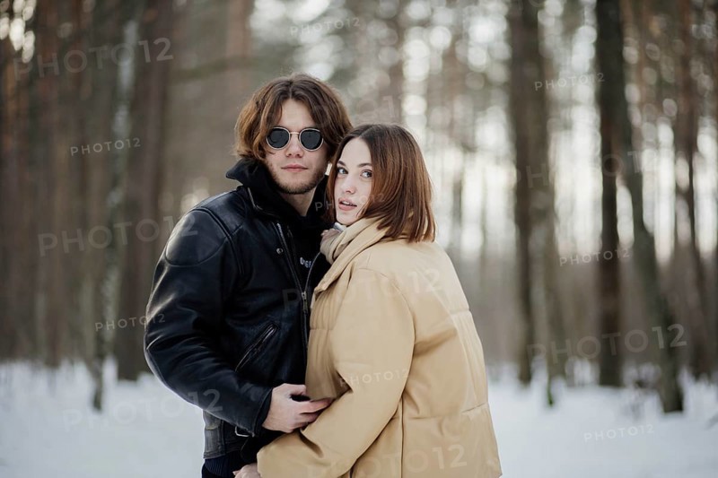 Couple poses in a snowy forest, looking at camera, dressed in winter jackets and scarves