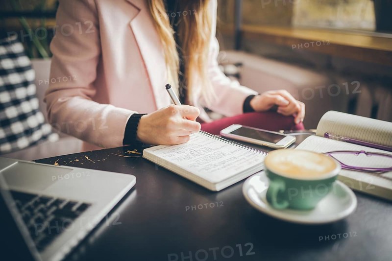Close up caucasian woman hand on wooden table inside cafe makes notes in notebook. Subject freelancer blogger journalist at work. Unrecognisable person. Technology phone and laptop with cup coffee