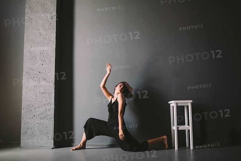 Social theme of female loneliness is pain suffering. Abastration of male domestic violence. Young beautiful Caucasian woman in black clothes, strange pose, white chair big dark room. Fear and despair