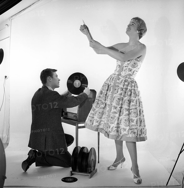 Gramophone in the 1950s. An man is kneeling by a combined recieving set and gramophone cabinet where he has opened the cover of the gramophone and holds a record in his hand.She is dressed in a typical 1950s dress looking happy. Sweden 1959. Kristoffersson ref CD65-2