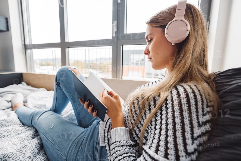 Blonde young caucasian woman lying on her dorm room bed next to a window putting on headphones, listening to music on a cold cloudy autumn day