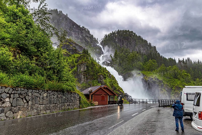 Woman tourist taking pictures of scenic double waterfall Lotefossen. Summer rainy day. The road passes through water jets. Cloud of water dust hangs in the air. Travel to Norway