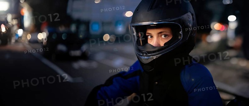 A person in a helmet and motorcycle gear standing by the street with urban lights in the background