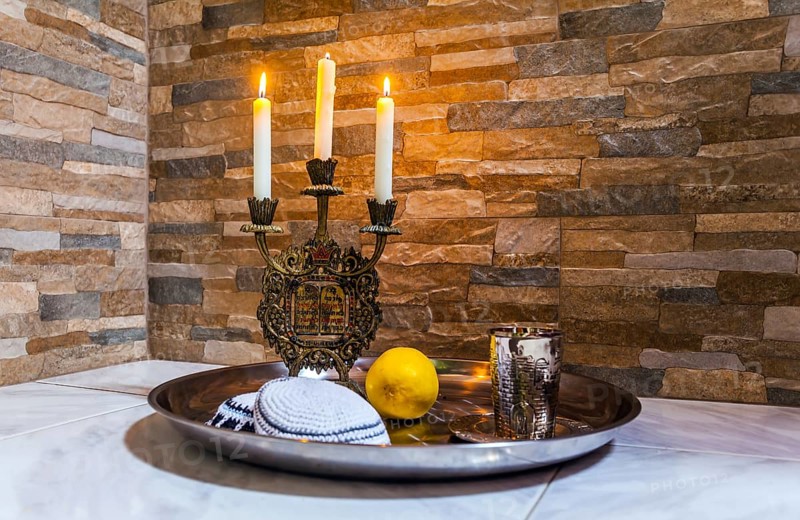 Beautiful candlestick with burning candles, a silver glass for blessing and a white yarmulke. Background stone wall. Still life with objects Judaica. Concept of artistic photography