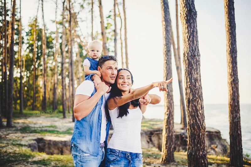 Happy young family of three people resting in a park outside the city. daughter sits at daddy on shoulders, and parents show direction forward by a hand for horizon.dressed in stylish denim clothes