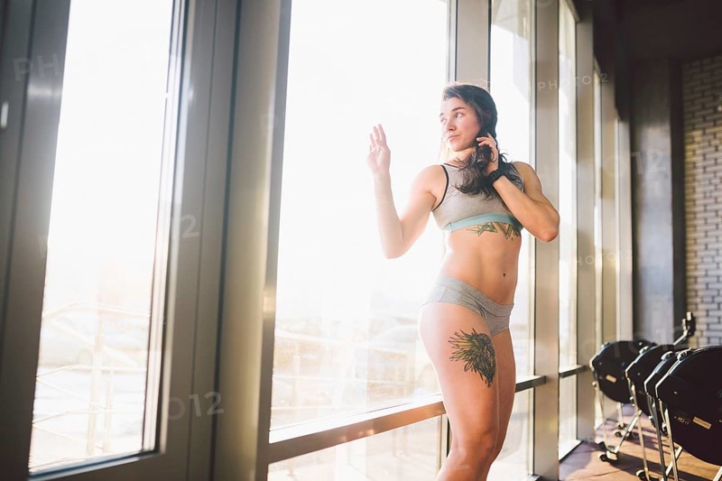 Young caucasian sporty woman with curly long hair poses in sportswear near a panorman window in a fitness club. Young female athlete model with beautiful body stands in the gym with a tattoo
