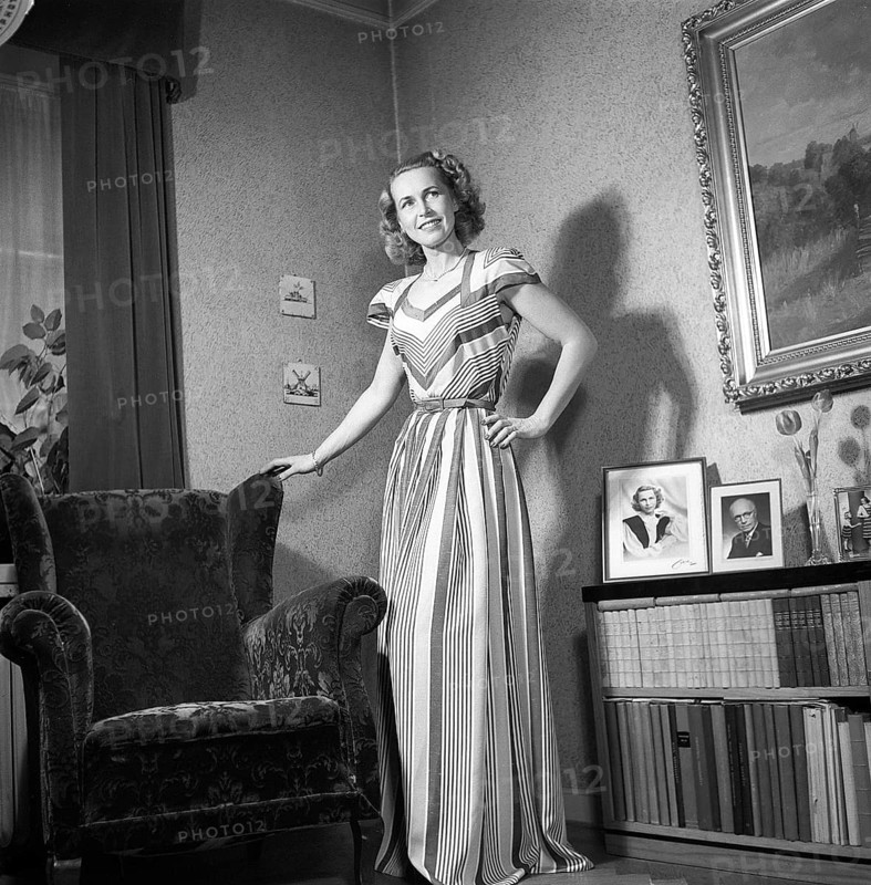 Fashion of 1946. A woman pictured wearing the typical fashion of the postwar era. She is wearing a long dress, the fabric of which is a pattern of narrow and wide fields of different colours. She is standing in a room by an armchair.Kristoffersson ref Y129-3