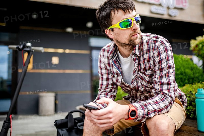 Individual urban environmental friendly transport electric kick scooter parked near man sitting on wooden bench in recreation area. Male use phone, surfing Internet, online shopping at smartphone