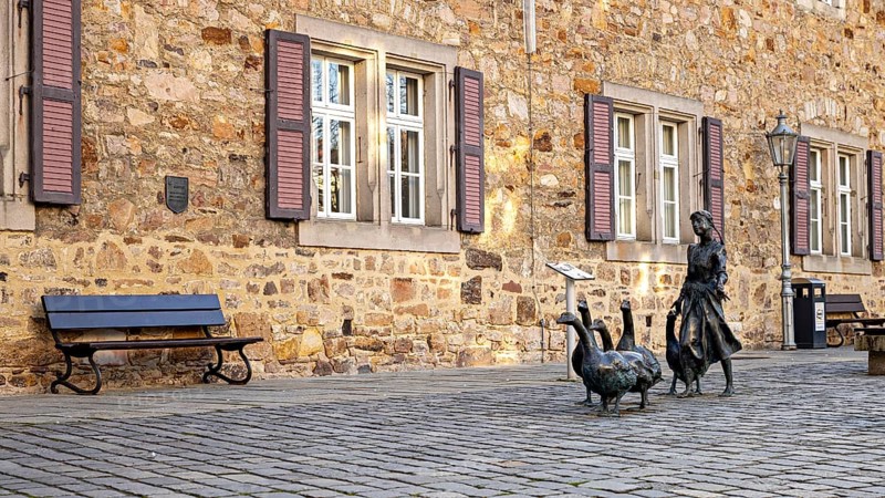 Scene in front of a stone building with shutters, a bench and a statue of a girl with geese, The Gänseliesel Monument in Melsungen