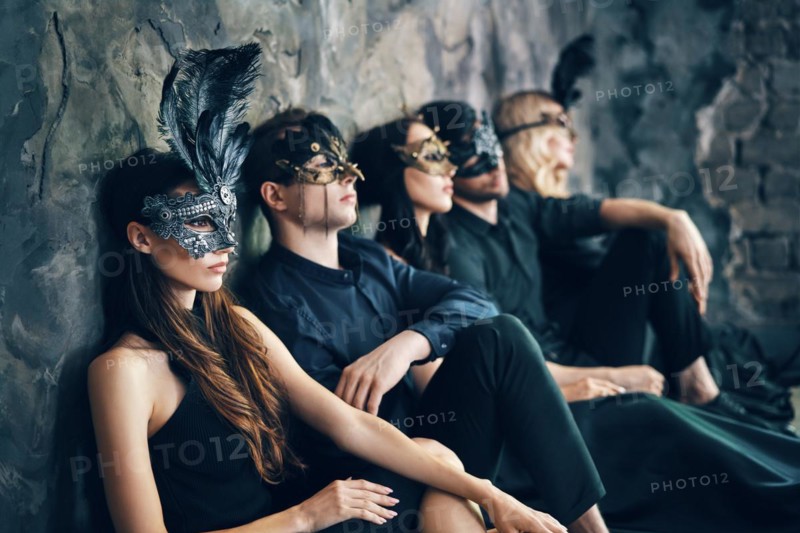 Group of friends in masquerade carnival mask sitting on floor relax after party. Beautiful women and men wearing venetian mask. Fashion, friends concept