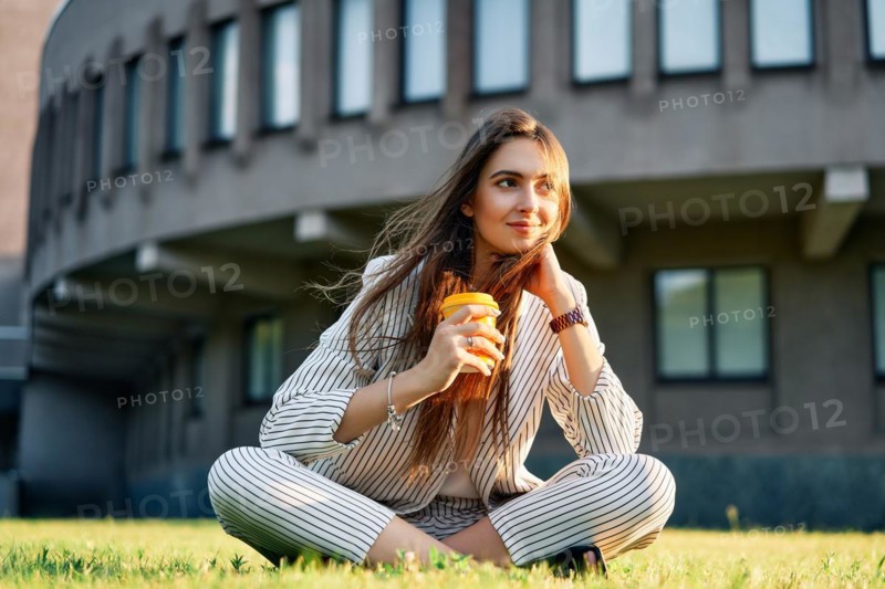 Young stylish woman drinking coffe and relax sitting on a grass in city park. Break time concept