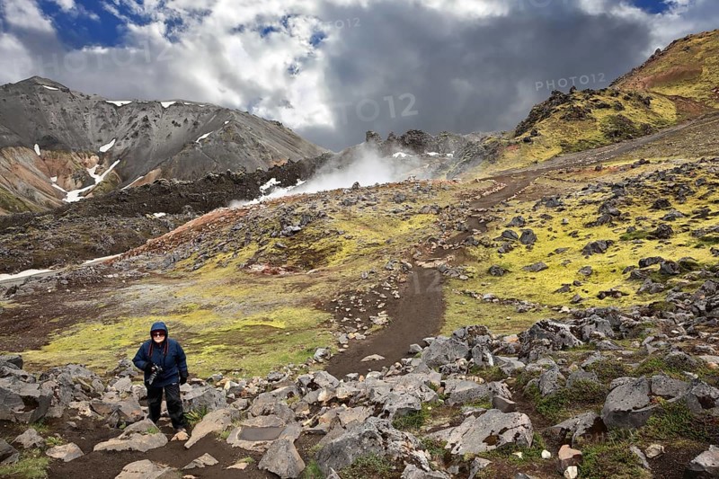 Rhyolite mountains smoldering underground heat. In the hollows lie unmelted snow patches from last year. Mature woman tourist wearing warm up the trail. Iceland in July