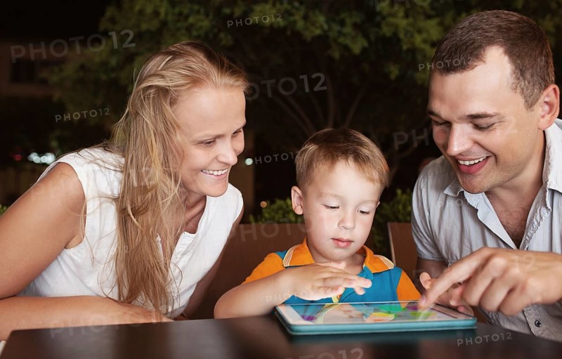 Young parents and little son in a cafe in the evening. Father and son using touch pad, mother watching them