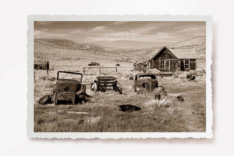 Several rusting car wrecks in a meadow in front of a wooden house in the former gold mining town of Bodie. Photo in sepia tone with white jagged edge, reclining shadow. Several rusting old car wrecks in a meadow in front of a wooden house in the former gold mining town of Bodie. Photo in sepia tone with white jagged edges