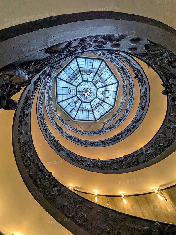 View from below of an ornate spiral staircase with a geometric skylight at the top, showcasing intricate metalwork. The Vatican