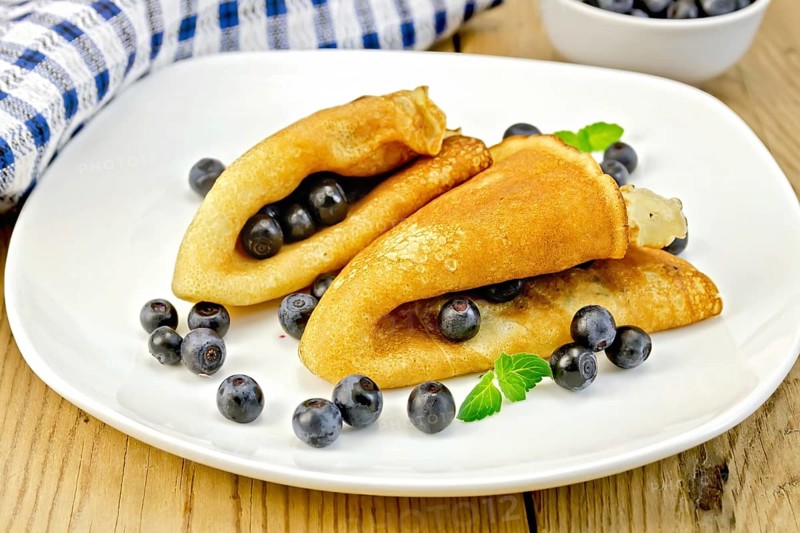 Two pancakes with blueberries and mint on a white plate, napkin, bowl on a wooden boards background