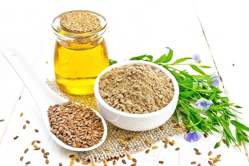 Flaxseed flour in a bowl, linenseeds in a spoon, oil in glass jar on burlap napkin, leaves and blue flaxen flowers on wooden board background