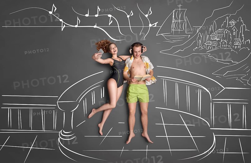 Love story concept of a romantic couple on the shore against chalk drawings background, view from above. Man wears headphones and reads a book, woman takes selfie with a smartphone