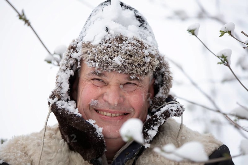 An elderly man in a snowy winter hat smiles and looks into the camera