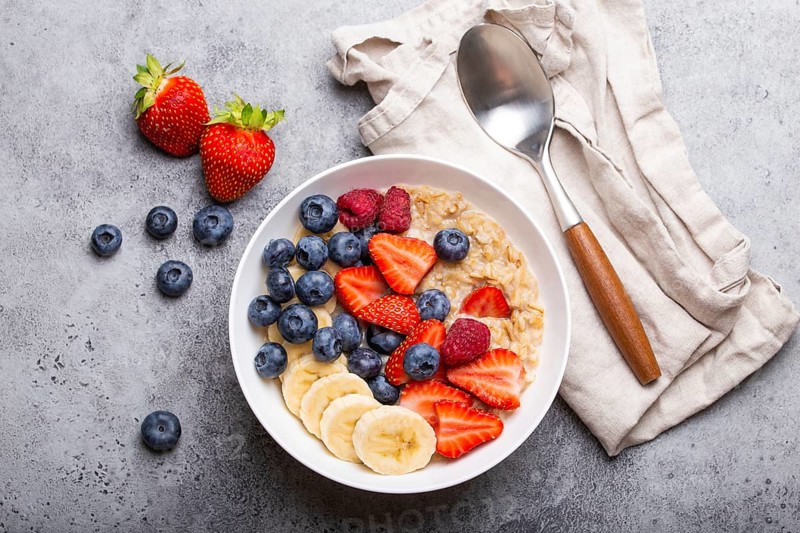 Oatmeal porridge with fruit and berries in bowl with spoon on white wooden background table top view, homemade healthy breakfast cereal with strawberry, banana, blueberry, raspberry, food photography