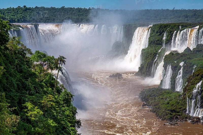 View to Devil's Throat and Iguazu Falls gorge with brown river, white cascading falls and lush green rainforest in sunshine