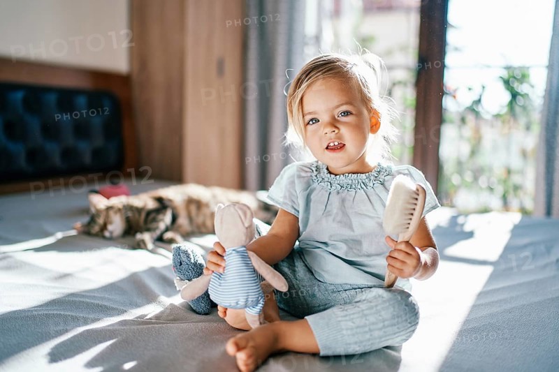 Little smiling girl sitting on the bed with a teddy bear and a comb in her hands.