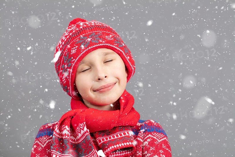 Dreamy child in warm clothing against a winter background. A boy with a red knitted hat and closed eyes. Fashionable boy in the studio in white snowflakes
