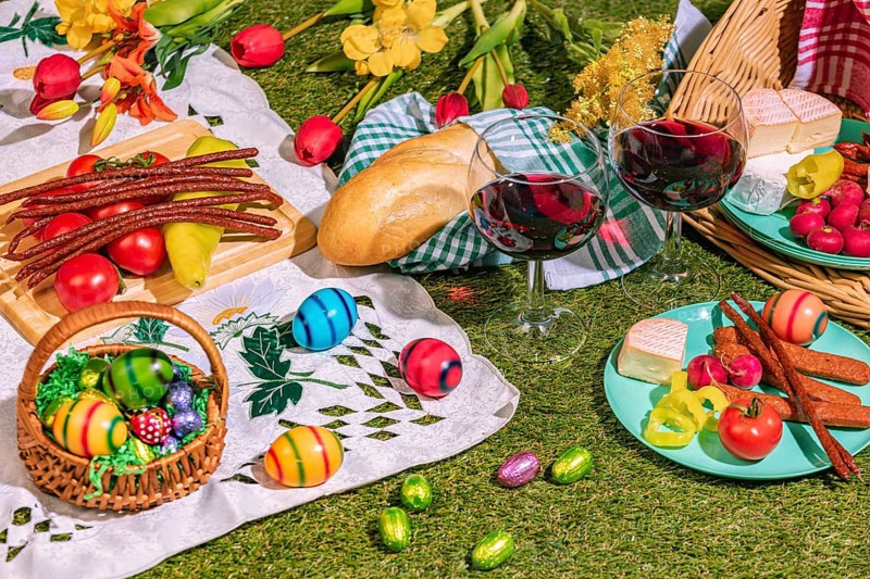 A picnic with two wine glasses on grass, surrounded by bread, sausage, cheese, vegetables and colourful Easter eggs
