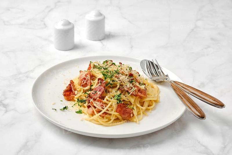 A delicious plate of spaghetti carbonara topped with crispy bacon, grated cheese, and fresh herbs on a marble table. Small salt and pepper shakers complete the casual dining setting