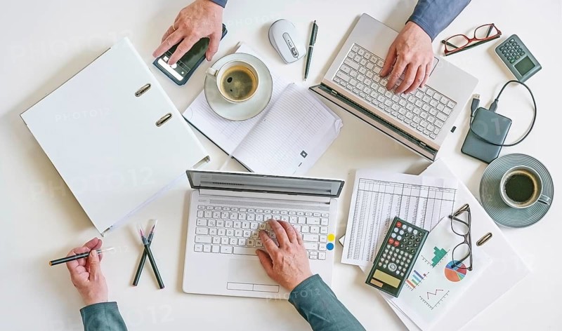 Top view of a white office desk with hands of two people working on laptops with papers, calculator, and coffee, teamwork and accounting concept, high angle shot from above, copy space, selected focus
