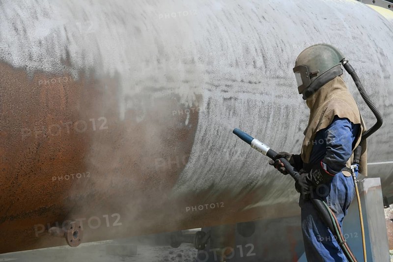 A man weating full safety gear sandblasts the steel casing of a meat drying machine for a rendering plant with ground glass
