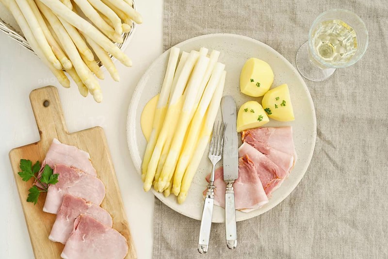 Traditional German dish with white asparagus, boiled ham, hollandaise sauce and potatoes, served on a light-coloured plate, accompanied by a glass of white wine