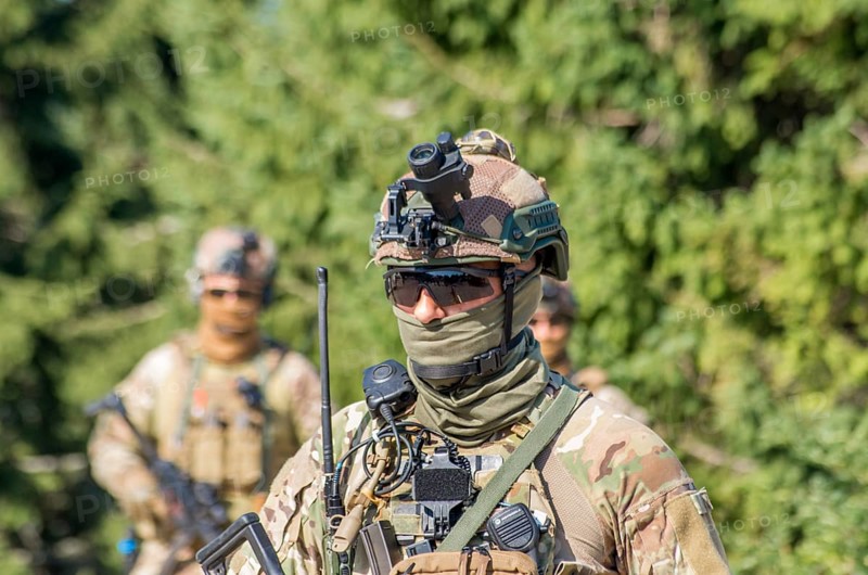 A soldier in camouflage uniform with tactical gear and sunglasses patrols a forest area