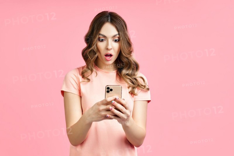 Portrait of a shocked pretty millennial hipster girl using mobile phone on pink background. surprised concept