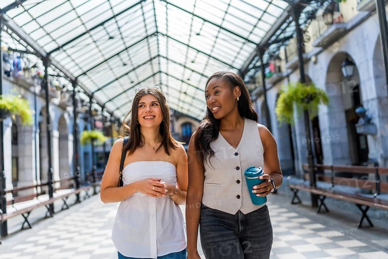 Two young businesswomen are walking and talking in a covered gallery, enjoying a break and holding reusable coffee cups