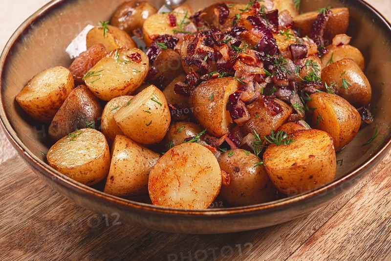A bowl filled with golden roasted potatoes seasoned with spices and garnished with caramelized onions and fresh herbs sits on a wooden table, showcasing a flavorful dish prepared for a meal