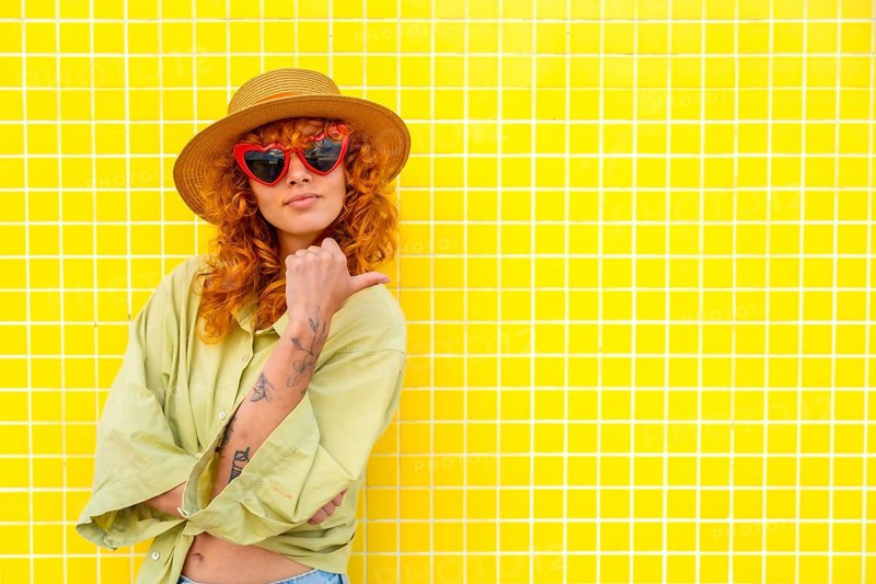 Fashionable influencer posing confidently against a vibrant yellow tiled wall, pointing with her thumb while showcasing her stylish look