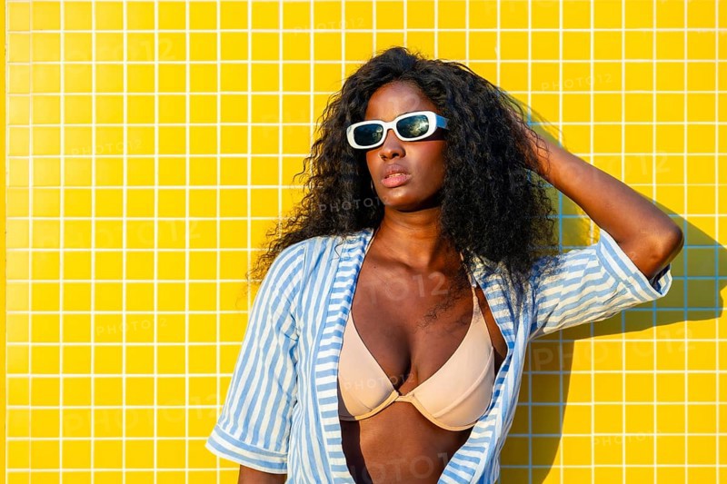 Stylish model with sunglasses and striped shirt posing against a vibrant yellow tiled wall, embodying summer fashion and confidence