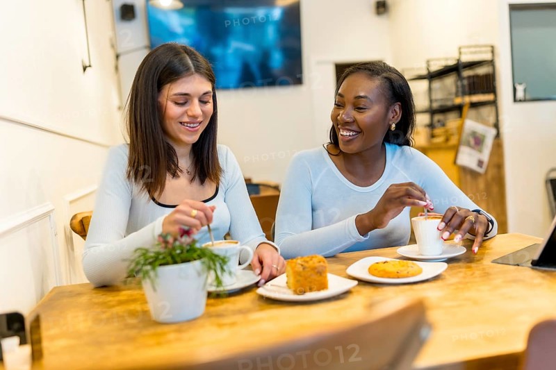 Two young women enjoying a coffee break, chatting and laughing together in a vibrant, modern cafe filled with natural light