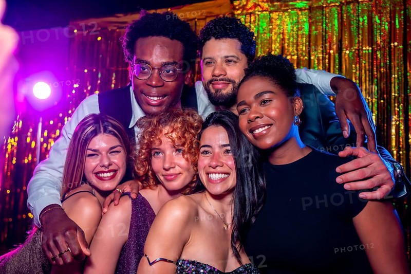 Group of cheerful elegant friends posing together for a photo at nightclub party with disco lights and gold tinsel curtain backdrop
