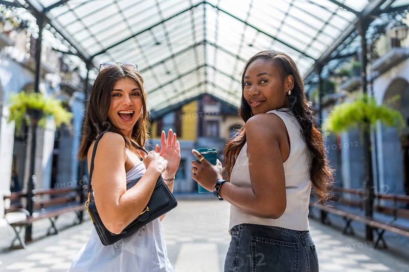 Two cheerful female friends walking away while waving and holding a reusable coffee cup in the city