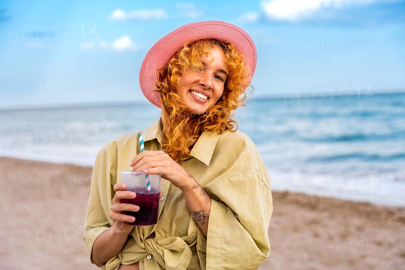 Young woman with curly red hair and pink hat enjoying a refreshing cold beverage on a sunny beach