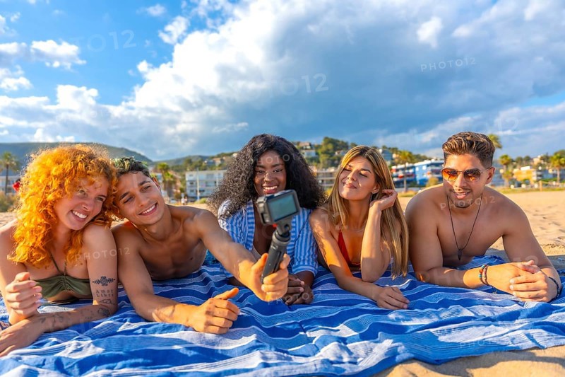 Happy multi ethnic friends lying together on a towel, capturing a joyful selfie on the beach while enjoying a sunny summer day