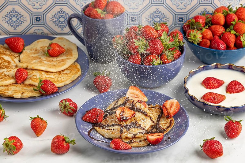 Icing sugar falls on pancakes with strawberries and chocolate syrup, fresh strawberries and yoghurt in the background