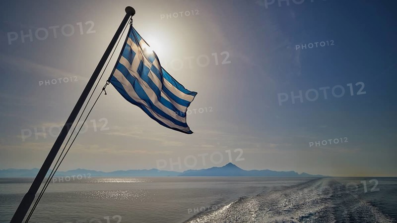 Greek flag in the foreground, over a widening sea under a blue sky, crossing Rhodes to Symi, ferry, Symi, Dodecanese, Greek Islands, Greece, Europe