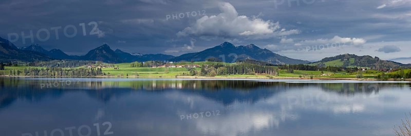 Panorama at sunrise over Lake Hopfensee to the sunlit western shore with farms, behind it receding thunderclouds, Lake Hopfensee, Ostallgäu, Allgäu, Bavaria, Germany, Europe