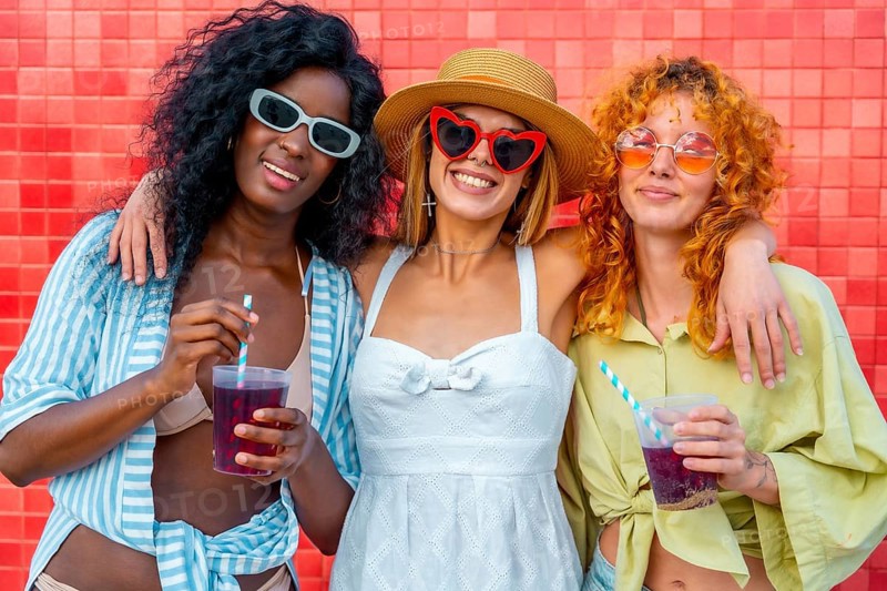 Three multi ethnic young women hugging each other and drinking juice with straw, standing against a red tiled wall